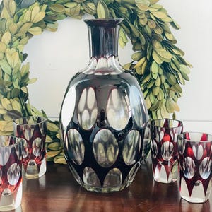 Puede incluir: Una jarra de vidrio decorativa y vasos de chupito a juego. La jarra es de color rojo oscuro con recortes ovalados, y los vasos tienen un diseño similar. Una corona verde está en el fondo. El conjunto se exhibe sobre una superficie de madera.