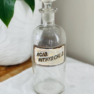 May include: A clear glass bottle with a glass stopper and a white label with black lettering that reads "ACID. NIT.HYD.CHL.D."