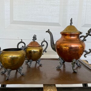 Può includere: Un set da tè in rame vintage composto da una teiera, un creamer e una zuccheriera. Ogni pezzo presenta manici decorati e arrotolati e poggia su tre piedini decorativi.