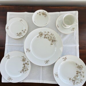 May include: A white porcelain dinnerware set with a floral design in shades of brown and gold. The set includes plates, bowls, and a teacup with a saucer. The edges of the plates and teacup have a gold trim.