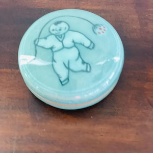 May include: A round, light teal-colored ceramic trinket box with a lid. The lid features a whimsical illustration of a child figure in a light blue outfit, holding a flower. The background is a dark brown wooden surface.