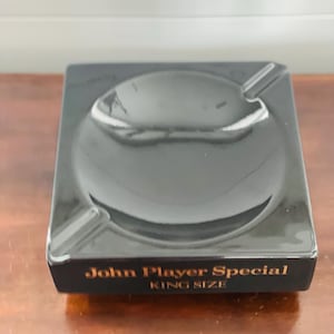 May include: A black ashtray with a rounded bowl and a rectangular base. The text "John Player Special" and "KING SIZE" are printed in gold lettering on the front. The ashtray is on a wooden surface.