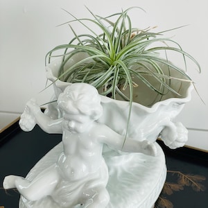 Puede incluir: Una maceta de cerámica blanca con un querubín que sostiene un arreglo floral. La maceta tiene un borde festoneado y contiene una planta verde. El querubín está en una pose reclinada, agregando un toque decorativo.