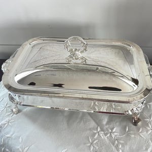 Può includere: Un piatto da portata rettangolare argentato con coperchio. Il piatto ha manici e piedini decorati e una maniglia decorativa sul coperchio. Il piatto è appoggiato su un panno bianco ricamato.