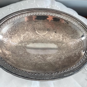 May include: Oval silver serving tray with ornate, engraved floral and scrollwork patterns. The tray has a detailed, raised border with a chain-like design. The interior of the tray reflects the surrounding environment.
