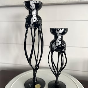Puede incluir: Dos candelabros de vidrio negro con diseños de salpicaduras blancas. El más alto tiene un tallo calado y delgado, mientras que el más corto tiene un diseño más compacto. Ambos tienen bases redondas y se exhiben sobre una superficie blanca.