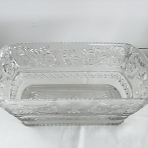 May include: Clear glass rectangular serving dish with ornate floral and scrollwork detailing. The dish has a slightly rounded shape and a textured rim. A decorative piece for serving food or displaying items.