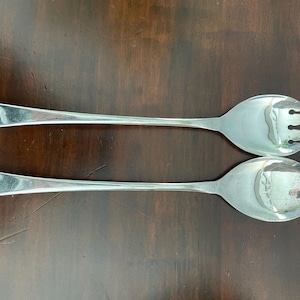Puede incluir: Un juego de dos utensilios para servir ensalada de plata. El tenedor tiene tres dientes y la cuchara tiene un cuenco redondeado.