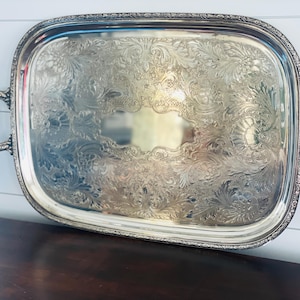 May include: A large, rectangular silver-toned serving tray with rounded corners and two ornate, curved handles. The surface features an intricate, embossed floral and scrollwork pattern, while the outer rim is decorated with a detailed border.