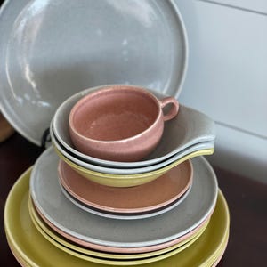 Könnte beinhalten: Ein Stapel vintage Keramikgeschirr in den Farben Grau, Rosa und Gelb. Das Set beinhaltet einen großen Speiseteller, einen kleineren Salatteller und eine Teetasse.