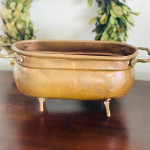 May include: A small, oval-shaped copper planter with two brass handles and three short legs. The planter is empty and has a slightly aged, rustic look.
