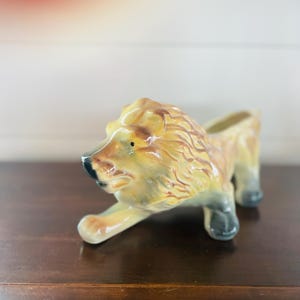 Puede incluir: Figura de león de cerámica en posición reclinada, con melena y cuerpo de color beige, y detalles negros en la nariz y las patas. El león tiene una pequeña abertura en la espalda, posiblemente para usar como maceta. Descansa sobre una superficie de madera oscura. Mide unos 20 cm.