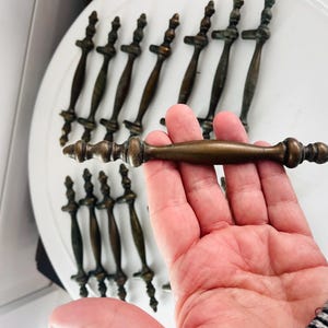 May include: Antique bronze cabinet pulls with decorative detailing. The pulls have a dark bronze finish and a classic design. The image shows several pulls, with one held in a hand for display. These pulls are suitable for furniture restoration.