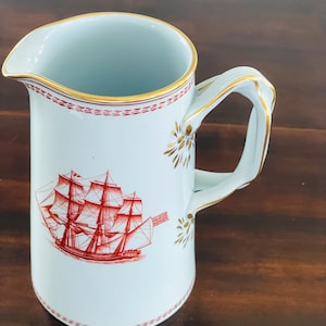 Puede incluir: Jarra de cerámica blanca con un diseño de barco rojo y dorado. La jarra tiene un pico y un asa con detalles dorados. El borde y la base presentan un patrón de puntos rojos. La ilustración del barco es detallada.