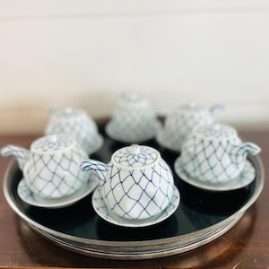 May include: A set of six small white porcelain teapots with matching saucers, each adorned with a blue crisscross pattern. The teapots are arranged on a silver-rimmed tray, creating an elegant presentation. The teapots are approximately 4 inches in height.