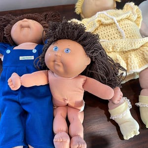 May include: Several vintage Cabbage Patch Kids dolls are displayed. One doll wears blue overalls, while another is dressed in a yellow crocheted dress. The dolls have soft bodies, plastic faces, and brown hair. The dolls are a nostalgic collectible.