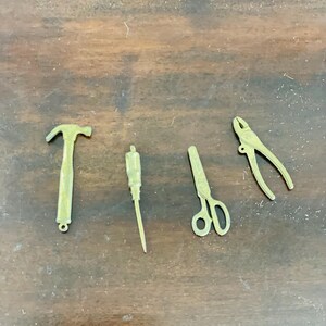 May include: Four small, gold-coloured tools are arranged on a wooden surface. The tools include a hammer, a screwdriver, scissors, and pliers. The tools are likely miniature or decorative items.