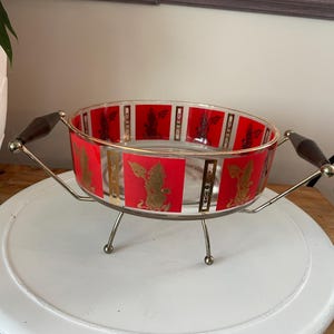 May include: A red and gold glass serving bowl with a metal stand and handles. The bowl has a geometric pattern with gold accents.