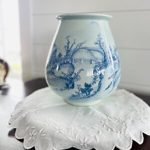 May include: A light blue ceramic vase with a rounded body and a flared rim, featuring a detailed blue transferware-style landscape illustration of a house, trees, and rolling hills encircling the exterior surface of the vessel.