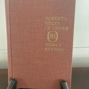Puede incluir: Un libro de tapa dura de color marrón rojizo apagado, titulado "Robert's Rules of Order". El título y el nombre del autor están en letras doradas. El libro se exhibe en un soporte negro.