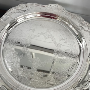 May include: A silver-coloured, ornate serving tray with a detailed floral pattern in the centre and along the scalloped edges. The tray has a reflective surface, and the design is classic and elegant.