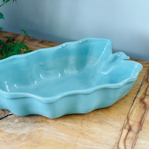 May include: A light blue ceramic baking dish shaped like a leaf. The dish is sitting on a wooden surface.