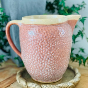 May include: A pink ceramic pitcher with a textured surface and a cream-colored rim. The pitcher is sitting on a gold-colored, rope-like coaster.