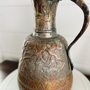 May include: An antique copper pitcher with an ornate design. The pitcher has a wide, round body and a long, curved handle. The surface is covered in intricate engravings of people and plants.