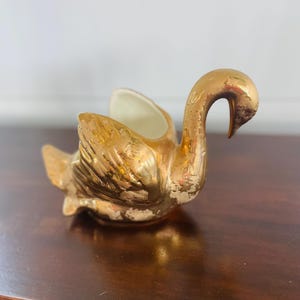 May include: A gold-colored swan figurine with a curved neck and outstretched wings. The ceramic swan has a distressed finish, revealing the white ceramic underneath. The swan is a decorative item, possibly a planter or a small dish.