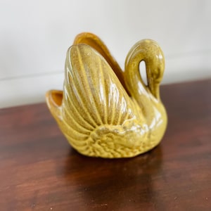 Könnte beinhalten: Eine goldgelbe Keramik-Schwanfigur. Der Schwan hat detaillierte Federstrukturen und einen gebogenen Hals. Die Figur ist wahrscheinlich ein Dekorationsartikel, möglicherweise für ein Zuhause oder als Sammlerstück. Der Schwan steht auf einer dunkelbraunen Oberfläche.