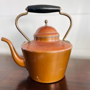 May include: A copper-colored teapot with a black handle and brass accents. The teapot has a rounded body, a curved spout, and a lid with a small knob. The handle is arched, and the teapot appears to be vintage.