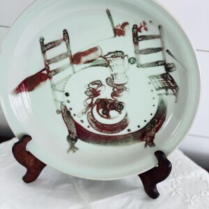Può includere: Un piatto rotondo in ceramica con smalto verde pallido caratterizzato da un'illustrazione dipinta a mano di una scena da pranzo. Il design raffigura un tavolo circolare con due sedie, una brocca e una ciotola di frutta in rosso scuro e verde.