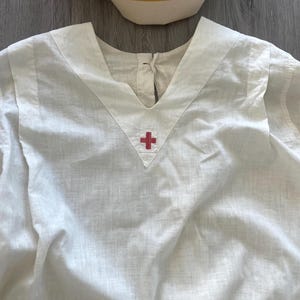 Op de afbeelding: Een witte tuniek met V-hals voorzien van een klein rood kruis-embleem in het midden van de halslijn en een bijpassende witte verpleegsterskap met een horizontale groene en gele streep langs de onderrand, gepresenteerd als een medisch uniform.