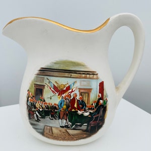 May include: White ceramic pitcher with a gold rim and a painted scene of the signing of the Declaration of Independence. The scene shows a group of men in period clothing gathered around a table, signing a document. The text "The Declaration of Independence, 1776" is printed below the scene.