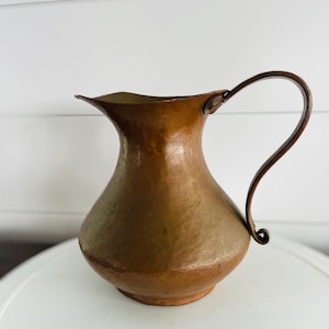 May include: A hammered copper pitcher with a curved handle. The pitcher has a wide mouth and a bulbous body. The handle is a darker brown color than the pitcher. The pitcher is sitting on a white surface.