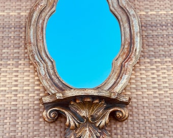May include: Ornate, antique-style wall mirror with a gold-toned frame and a blue reflective surface. The mirror features a decorative shelf and a detailed, leaf-like design at the bottom. The frame has a distressed finish, adding to its vintage appeal.