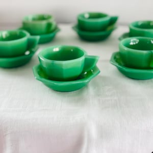 May include: A collection of six jadeite green teacups and saucers. The cups and saucers have a faceted design. The teacups have a handle and sit on matching, octagonal saucers. The set is arranged on a white tablecloth.