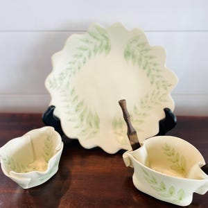 May include: A cream-colored serving set featuring a large scalloped plate and two smaller bowls, all adorned with a delicate green leaf pattern. A wooden utensil rests in one of the bowls. The set is displayed on a dark wooden surface.