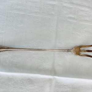 Könnte beinhalten: Eine lange, silberne Serviergabel mit drei Zinken. Der Griff hat ein verziertes, florales Design und einen Perlenrand. Die Gabel ist wahrscheinlich Vintage und liegt auf einer weißen Tischdecke.