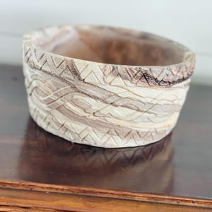 May include: A handcrafted, round wooden bowl with a textured, carved design. The bowl features a light brown and beige color palette with a decorative pattern around the exterior. The bowl is sitting on a dark brown surface.