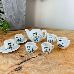 May include: A white porcelain tea set with a whimsical design featuring a cartoon character. The set includes a teapot, creamer, sugar bowl, and four teacups and saucers. Each piece is decorated with a blue and yellow illustration of a cartoon character holding a flower.