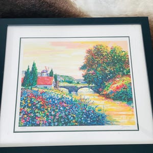 May include: Framed art print depicting a landscape with a bridge, river, and windmill. The artwork features vibrant flowers, trees, and a pastel sky. The print is set in a dark green frame, ideal for home decor.