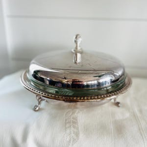 Può includere: Burriera argentata con inserto in vetro e coperchio a cupola. Il piatto ha un bordo decorativo con perline e poggia su piedini piccoli e ornati. Il coperchio presenta una piccola maniglia. La burriera è su una tovaglia bianca.