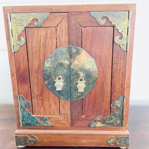 May include: A small, wooden cabinet with a reddish-brown finish. The cabinet features ornate brass corner brackets and a central decorative plate. The doors have a central brass handle with a floral design. The cabinet is a decorative storage piece.
