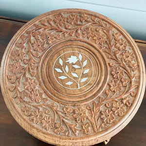 May include: A round, wooden decorative tray with intricate floral carvings. The tray features a central design with a floral and leaf pattern in white. The wood has a warm, natural tone, and the carvings add texture.