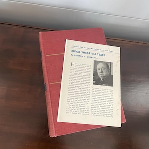 Może przedstawiać: Czerwona książka w twardej oprawie i zadrukowana strona z tytułem "Blood Sweat and Tears" autorstwa Winstona S. Churchilla. Strona zawiera czarno-biały portret Churchilla i tekst. Książka leży na ciemnej drewnianej powierzchni.