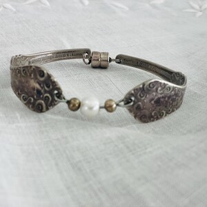 Può includere: Un braccialetto d'argento realizzato con cucchiai riciclati. Il braccialetto presenta due teste di cucchiaio con disegni ornati, collegate da una fila di perline e una chiusura magnetica. Una perla e due perline dorate sono al centro.