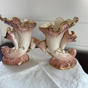 May include: A pair of cream and soft pink ceramic cornucopia vases featuring ornate, ruffled edges and a base decorated with raised, swirling patterns. Each vase is accented with metallic gold trim along the rims and the embossed details.
