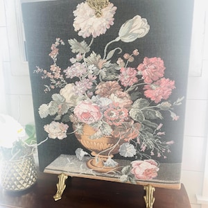 May include: A decorative tapestry screen featuring a floral arrangement in a golden urn against a dark background. The flowers are in shades of pink, peach, and white. The screen has brass feet and a decorative top.