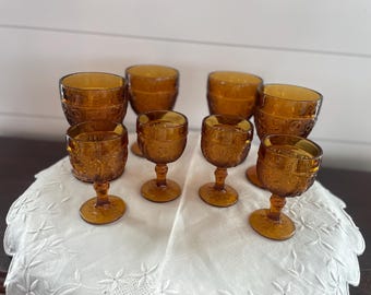 May include: A collection of seven amber-colored glass goblets and smaller stemmed glasses. Each piece features a textured, embossed floral pattern across the surface, with a round base, a short stem, and a wide, rounded bowl for each vessel.
